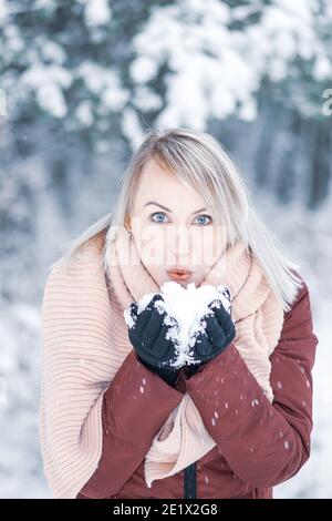 Donna bionda con cappotto marrone e sciarpa rosa che soffia la neve dalle mani con guanti neri durante l'inverno sfondo bianco della foresta Foto Stock