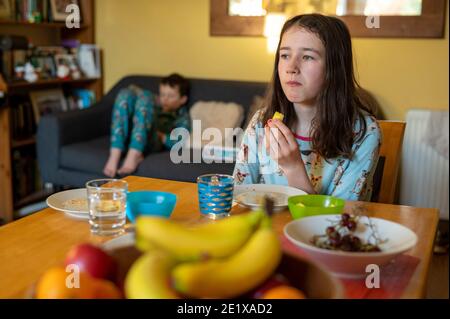 Edimburgo, Scozia, Regno Unito. Gennaio 2021. Una ragazza di 14 anni che mangia frutta al tavolo della colazione con il fratello minore distratto in background Foto Stock
