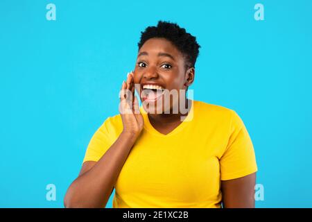Black Plus-Size Donna gridante tenendo la mano vicino alla bocca, sfondo blu Foto Stock