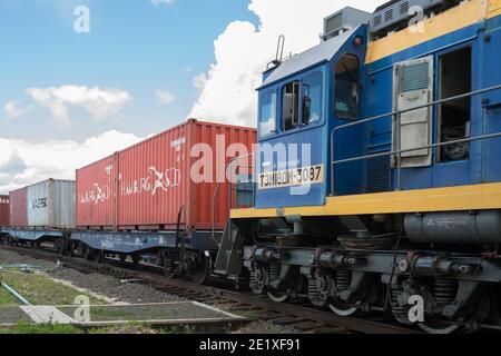 Una locomotiva con un conducente in cabina come parte di un treno merci con container si trova sulla pista ferroviaria in una giornata estiva soleggiata. Foto Stock