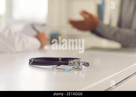 Primo piano di stetoscopio terapeuta sul tavolo sopra gli uomini medico e. pazienti che parlano in background Foto Stock