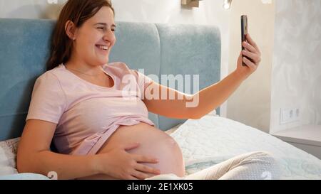 Donna incinta sorridente in attesa del bambino seduto a letto e chiamando un amico o un medico in videochiamata in conferenza. Donna incinta che usa lo smartphone Foto Stock