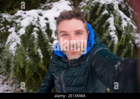 giovane felice e attraente uomo turistico che prende ritratto selfie con Telefono cellulare allegro nel parco della città durante le nevicate di Natale godendo e giocando w Foto Stock