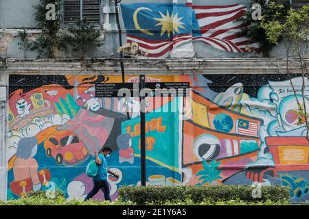 Un uomo che indossa una maschera facciale come precauzione contro la diffusione di Covid-19 cammina oltre un murale sulla strada a Kuala Lumpur. Foto Stock