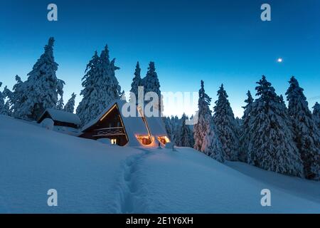 Fantastico paesaggio invernale con casa in legno con finestre in montagne innevate e cielo notturno. Vacanza di Natale e concetto di vacanza invernale Foto Stock