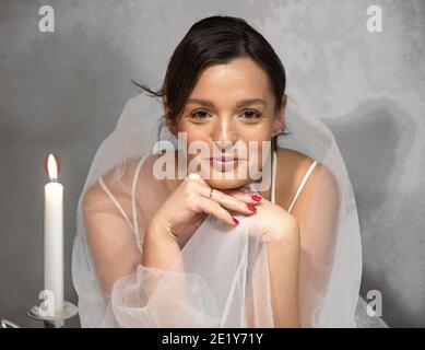 Elegante donna a lume di candela, drappeggiata in un morbido velo bianco, che irradia serenità e grazia in un ritratto intimo e romantico Foto Stock