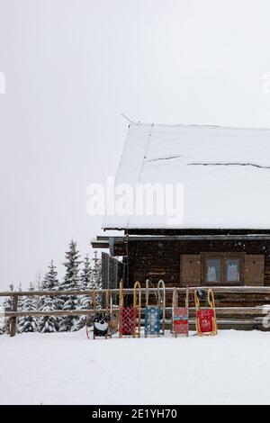 Colorate slitte in legno appoggiate a una recinzione in legno di fronte a una cabina innevata. Alberi e terreno ricoperti di neve, e cieli bianchi con le nuvole. Foto Stock