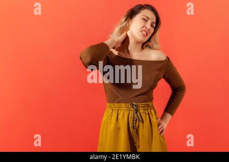 Dolore al collo o dolore. Ritratto di bella blonde giovane donna adulta in blusa marrone in piedi e tenendo il suo collo doloroso. Studio interno con isol di ripresa Foto Stock