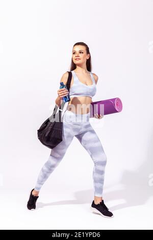 Donna sportiva sicura di sé in abbigliamento sportivo bianco con borsa, tappetino fitness e bottiglia con acqua che si fa male in palestra per allenarsi, pompando i muscoli. Interni Foto Stock