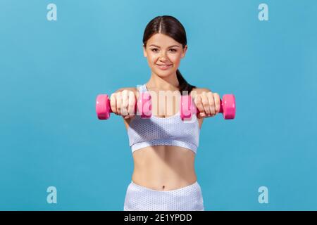 Sorridente donna atletica sana in sportswear che tiene i manubri nelle mani, mostrando il suo abs, facendo esercizi per mantenere una buona forma del corpo. Riprese in studio al coperto Foto Stock