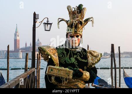 Uomo vestito in costume tradizionale decorato e maschera dipinta durante il Carnevale di Venezia con alle spalle il Monastero di San Giorgio, Venezia, Italia Foto Stock