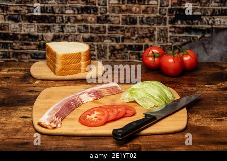 Pancetta, lattuga e pomodoro su un tagliere con un coltello pronto per preparare un panino BLT. Foto Stock