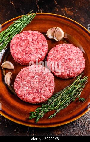 Carne di hamburger cruda tagliata con carne di manzo macinato ed erbe su un piatto. Sfondo scuro. Vista dall'alto Foto Stock