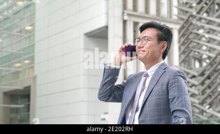 Uomo d'affari asiatico con cravatta e giacca, sorridente e parlando al telefono con edifici moderni sullo sfondo. Foto Stock