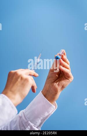Primo piano sulla mano di sconosciuta donna caucasica medico tenuta siringa con ago e ampolla di vaccino di fronte parete blu - covid-19 coronavir Foto Stock