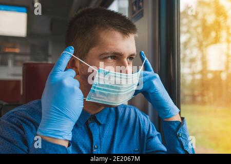 Giovane ragazzo caucasico in guanti protettivi mette su una maschera medica in un treno elettrico moderno e guarda fuori la finestra. Foto Stock