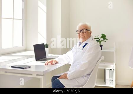 Medico professionista senior positivo in uniforme seduto con il computer portatile in ufficio medico e guardando la fotocamera Foto Stock