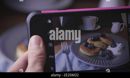 Per fare una foto con smartphone su frittelle di mirtillo in un piatto. Cucina tradizionale per colazione o brunch. Frittelle dolci fatte in casa, tazza di caffè, spo Foto Stock