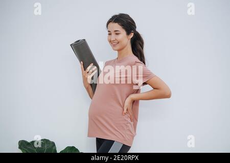 Donna incinta felice al club di yoga, tenendo il tappeto arrotolato, in posa per una foto Foto Stock