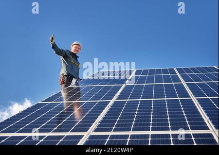 Lavoratore maschile dando pollici e sorridendo mentre si monta il pannello solare fotovoltaico blu sotto il bel cielo nuvoloso. Concetto di fonti energetiche alternative e innovazioni. Foto Stock