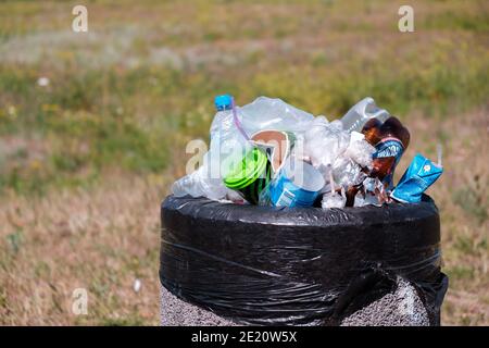 KRYVYI RIH, UCRAINA - AGOSTO, 2020. Bottiglie e sacchetti di plastica in bidone per rifiuti sovraccaricato su un'erba nel parco cittadino Foto Stock