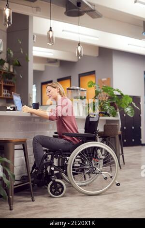 Donna d'affari in sedia a rotelle che lavora su laptop in cucina Ufficio moderno occupato Foto Stock