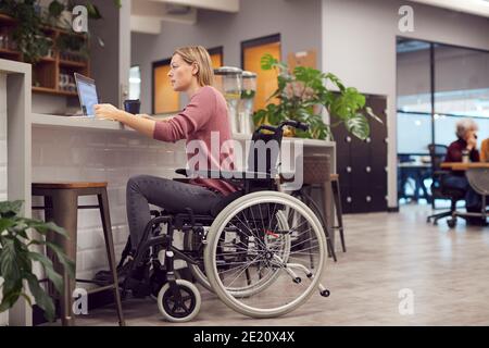 Donna d'affari in sedia a rotelle che lavora su laptop in cucina Ufficio moderno occupato Foto Stock