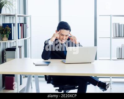 Bell'uomo d'affari asiatico in tuta concentrarsi guardando su schermo e. pensando con il dito che punta sulla testa mentre lavora con computer portatile acceso Foto Stock
