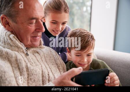 Nipoti sul divano a casa mostrando nonno come usare Telefono cellulare Foto Stock