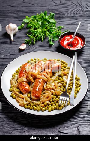 Piatto per la colazione con salsiccia di maiale, piselli verdi con sugo di cipolla e ketchup serviti con posate su sfondo di legno scuro, vista dall'alto, primo piano Foto Stock