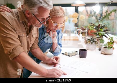 Coppia matura che esamina e firma finanze nazionali e documenti di investimento In cucina a casa Foto Stock