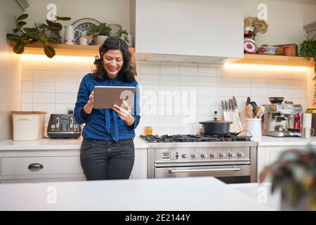 Donna matura a casa in cucina guardando Digital Tablet Foto Stock