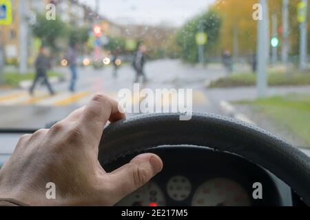 vista della mano del conducente sul volante di una macchina che si è fermata e passa pedoni a a. attraversamento pedonale in tempo di pioggia nuvoloso Foto Stock