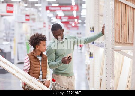 Vita ritratto di padre afroamericano e figlio shopping insieme nel negozio di hardware e utilizzando smartphone mentre si scelgono le tavole di legno per im. casa Foto Stock