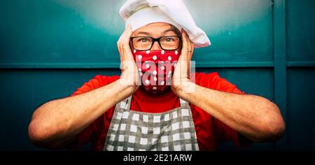 Shock e pazzo espressione spaventato uomo con cappello chef e. maschera medica di protezione rossa - coronavirus covid-19 economy restrizioni immagine concetto con peo Foto Stock