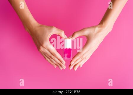 vista ritagliata della donna che fa il simbolo del cuore con le mani mentre tenendo una bottiglia di smalto per unghie su sfondo rosa Foto Stock