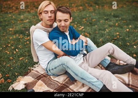 Coppia gay amorevole che ha date romantiche all'aperto. Due bei uomini seduti insieme sulla coperta nel parco e abbracciando. Concetto LGBT. Foto Stock