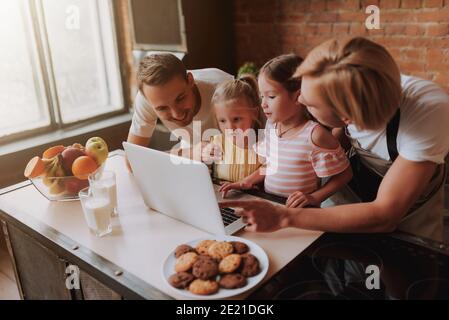 Coppia gay con le loro figlie adorabili che leggono la ricetta sul computer portatile e cucinando in cucina. Famiglia LGBT a casa. Foto Stock