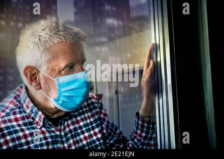l'uomo anziano in una maschera medica si trova davanti alla finestra e guarda la strada Foto Stock