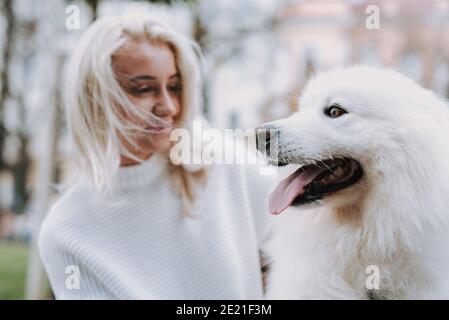 Giovane donna bionda sorridente che cammina il suo cane bianco soffice razza samoyed nel parco. Proprietario e animale domestico Foto Stock