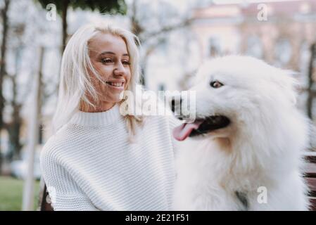 Giovane donna bionda sorridente che cammina il suo puppy bianco soffice razza samoyed nel parco. Proprietario e animale domestico Foto Stock