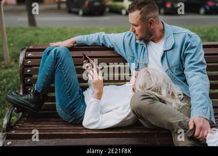 Donna bionda felice che guarda il video sul telefono cellulare mentre si riposa con il suo ragazzo su una panchina vicolo. Data romantica Foto Stock