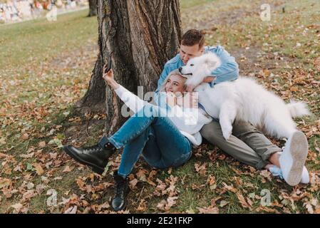 Giovane coppia sorridente con il cane seduto nel parco mentre creazione di foto sullo smartphone Foto Stock