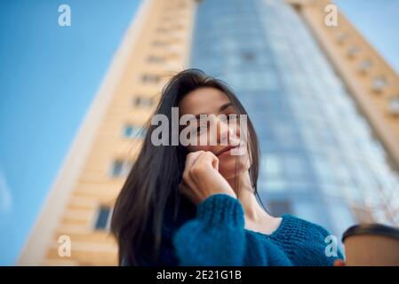 Primo piano di una donna dai capelli lunghi che parla dal cellulare sulla strada in un buon umore, alta torre dell'ufficio sta vedendo su sfondo sfocato Foto Stock