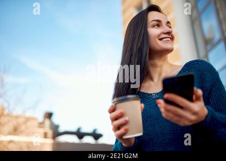 Giovane donna con caffè e smartphone in piedi vicino all'alto torre moderna in un quartiere degli affari e godendo del paesaggio urbano e. bel tempo Foto Stock