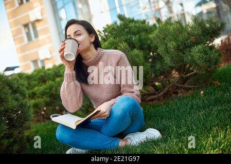 Donna giovane graziosa con un libro da giorno spesso sulle sue ginocchia sorseggiando una bevanda calda su un prato verde vicino all'alto e moderno edificio Foto Stock