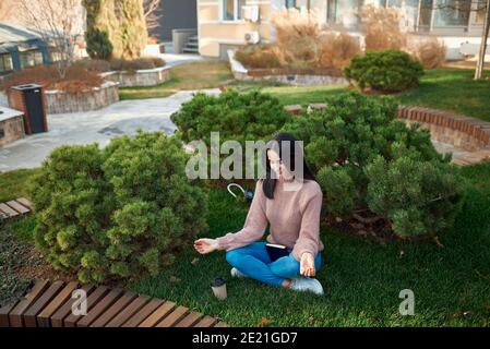 Vista ad alto angolo di una piacevole giovane femmina in cui rilassarsi una posa meditativa su un'erba in cortile accogliente dentro di fronte a edifici moderni Foto Stock