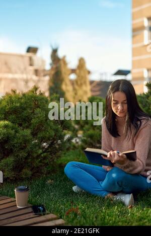 Giovane donna pusiva che gode di un libro interessante mentre si siede sull'erba in una facciata prima di un alto edificio moderno Foto Stock