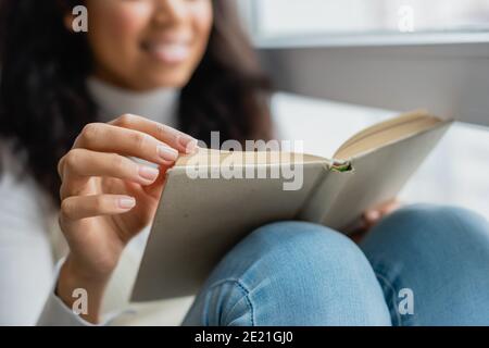 vista ritagliata del libro di lettura della donna afroamericana a casa, sfondo offuscato Foto Stock