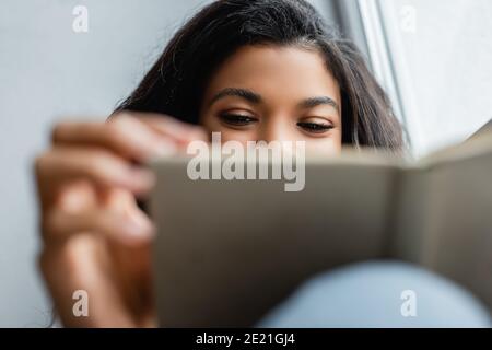 vista ritagliata del libro di lettura della donna afroamericana a casa, primo piano offuscato Foto Stock